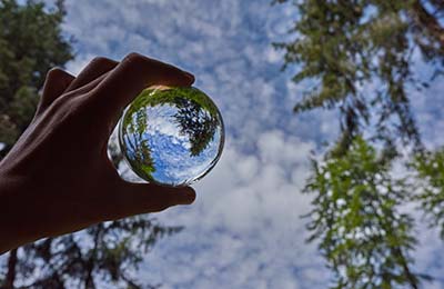 Photo of a person holding a clear glass marble with the blue sky and trees reflected